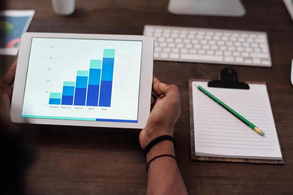 A person holding a tablet that shows a bar chart, beside a keyboard and a clipboard