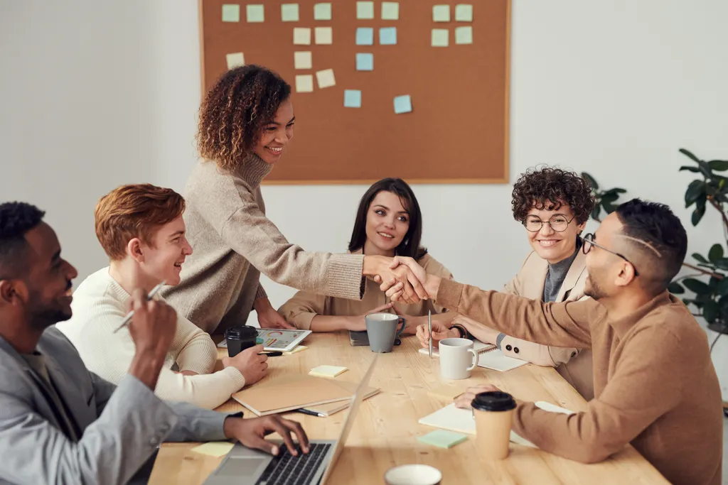 A team around a meeting table, two colleagues shaking hands across laptops and notebooks