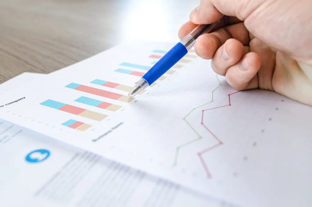 A hand holding a pen over printed bar charts and line graphs on a desk