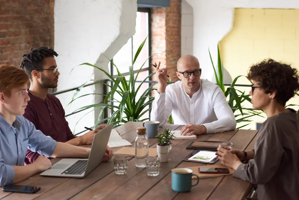 Four colleagues in discussion around a wooden table in a bright, plant-filled office