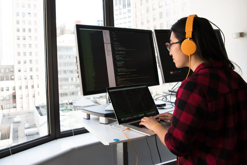 A developer wearing headphones working on code across two monitors by a city window